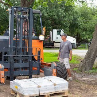 Man watches a forklift load pallet of gray pavers in front of a truck and trees on a sunny day.