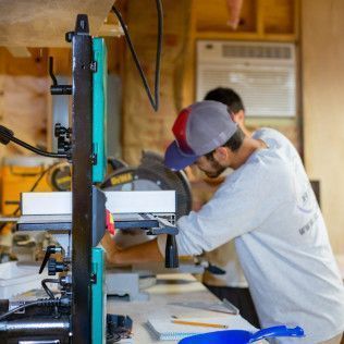 Two men work at a saw in a workshop, one wearing a hat.