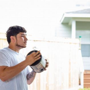 Man holding a paint can outdoors, looking left, wearing a white shirt near a fence and house.