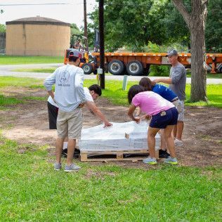 A group of people loading materials from a pallet, outdoors. A semi-truck is in the background.
