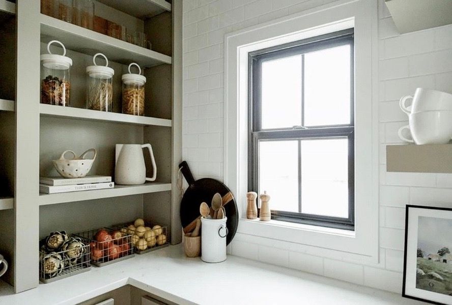 Kitchen with open shelves, window, and counter. Beige shelves hold jars, teapot, bowls. Window has black frame.