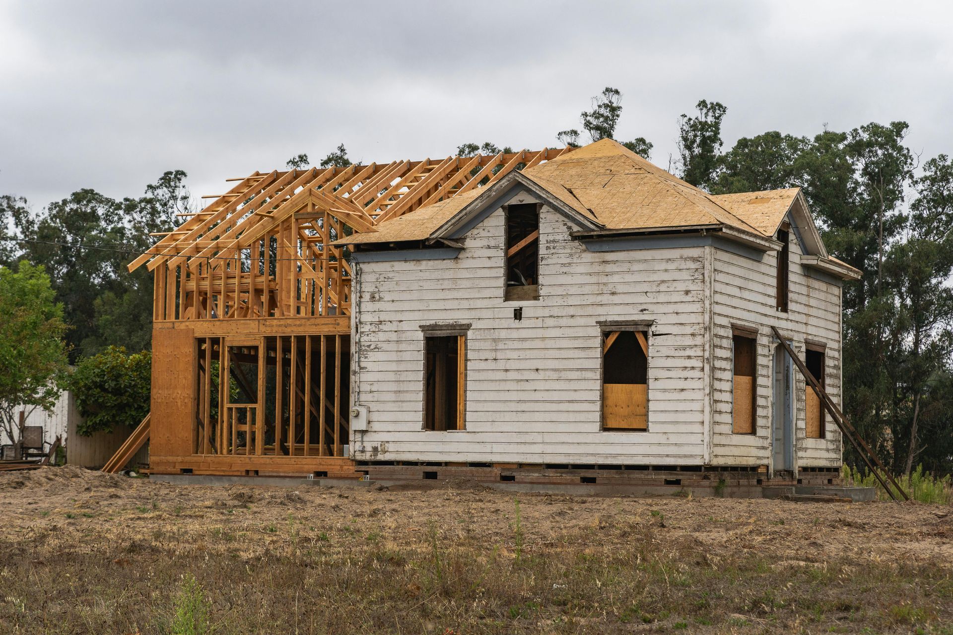Two-story house under renovation with exposed wood framing and weathered white siding, on a cloudy day.