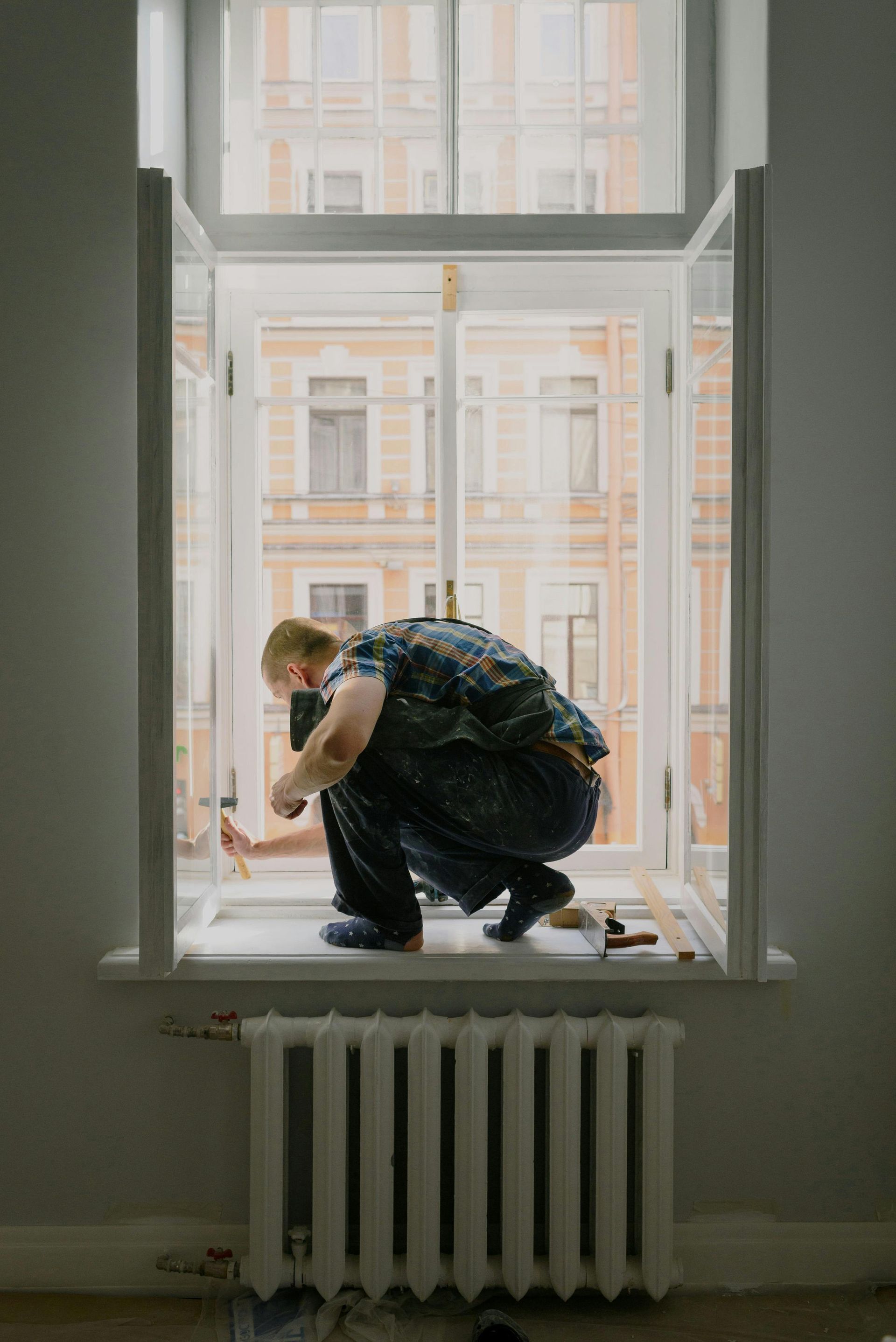 Man crouching on a windowsill, working inside. Buildings visible through the window.