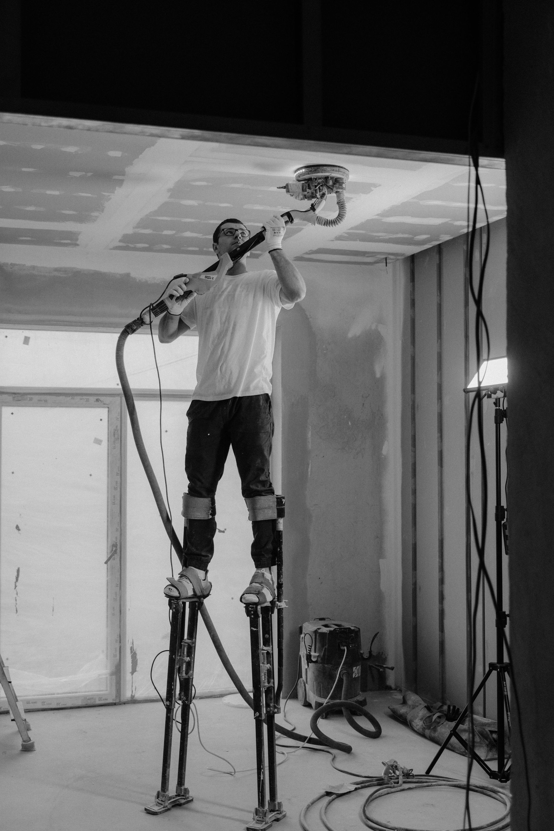 Man sanding ceiling with power sander while standing on stilts indoors.
