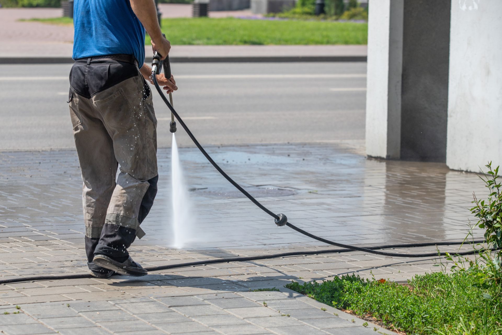 Person pressure washing a sidewalk next to a road, using a black hose.