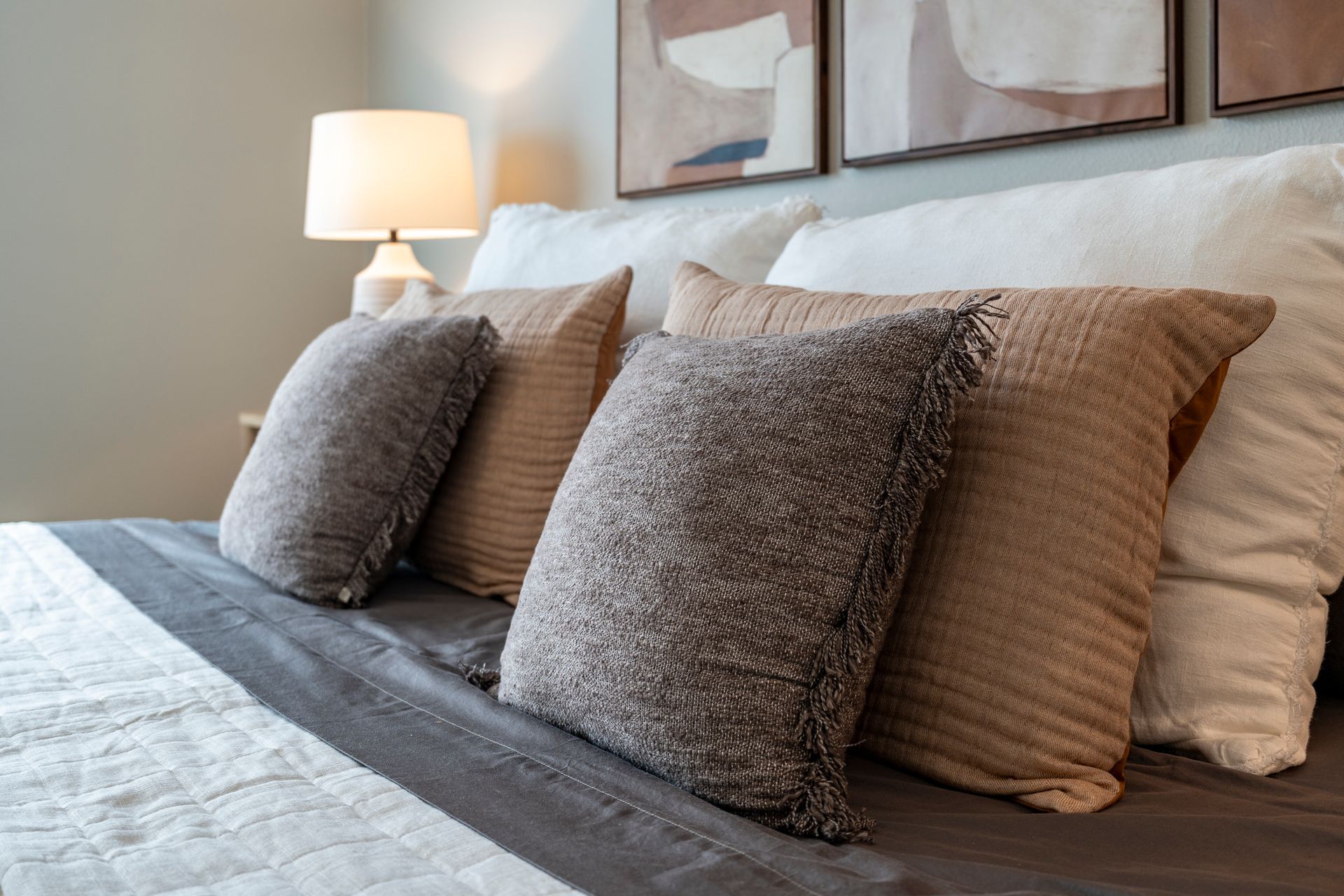 Bed with decorative pillows in neutral tones; a lit lamp sits beside the bed under framed art.