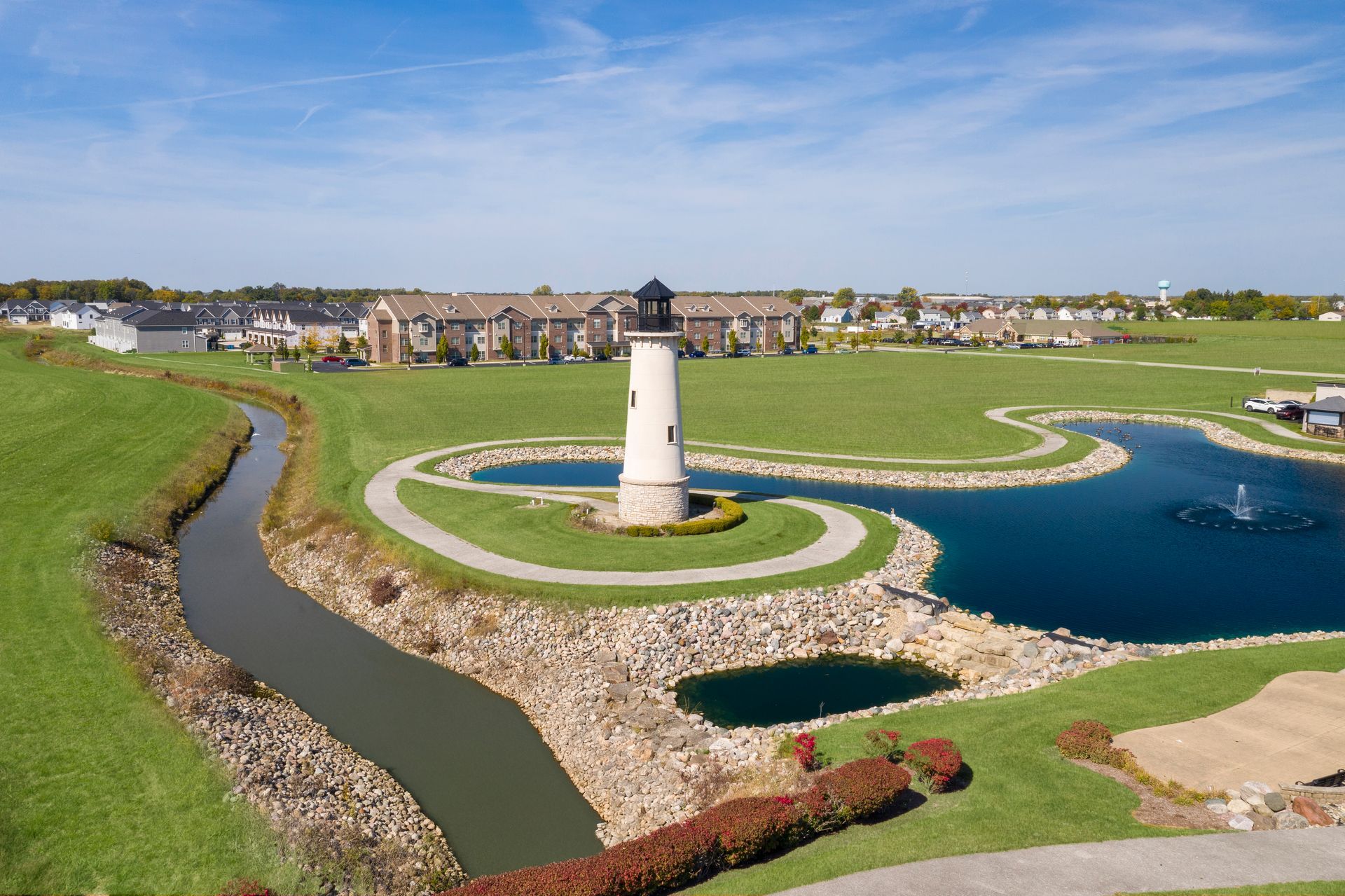 Lighthouse in a park with water features, surrounded by green grass and residential buildings on a sunny day.