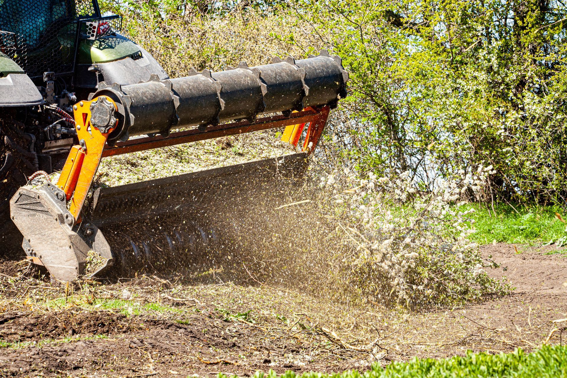 Tractor mulching vegetation, ejecting debris, in a field with green grass and foliage.