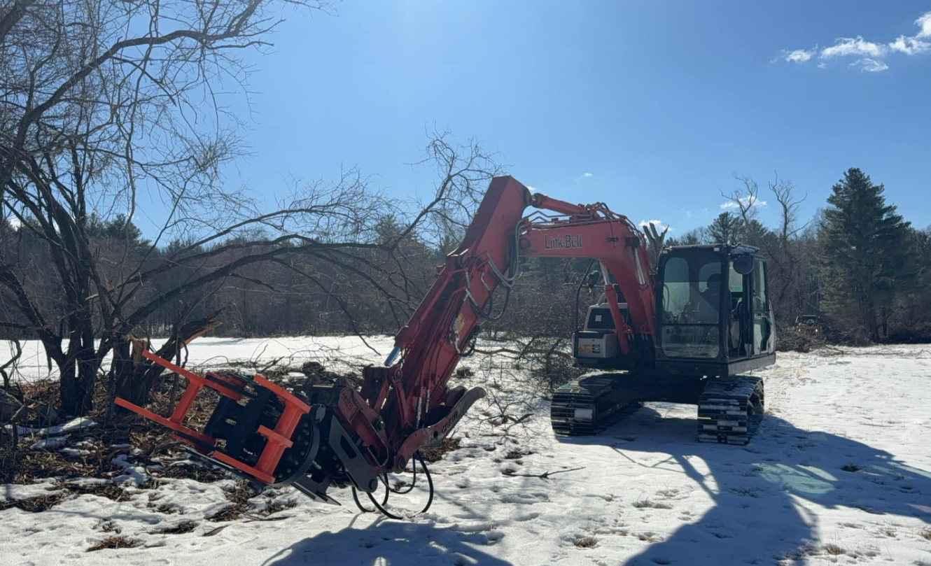 Red excavator clearing brush in a snowy field on a sunny day.