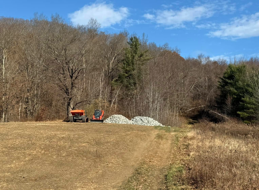 Tractor and mini excavator next to a pile of rocks in a field near a wooded area on a sunny day.