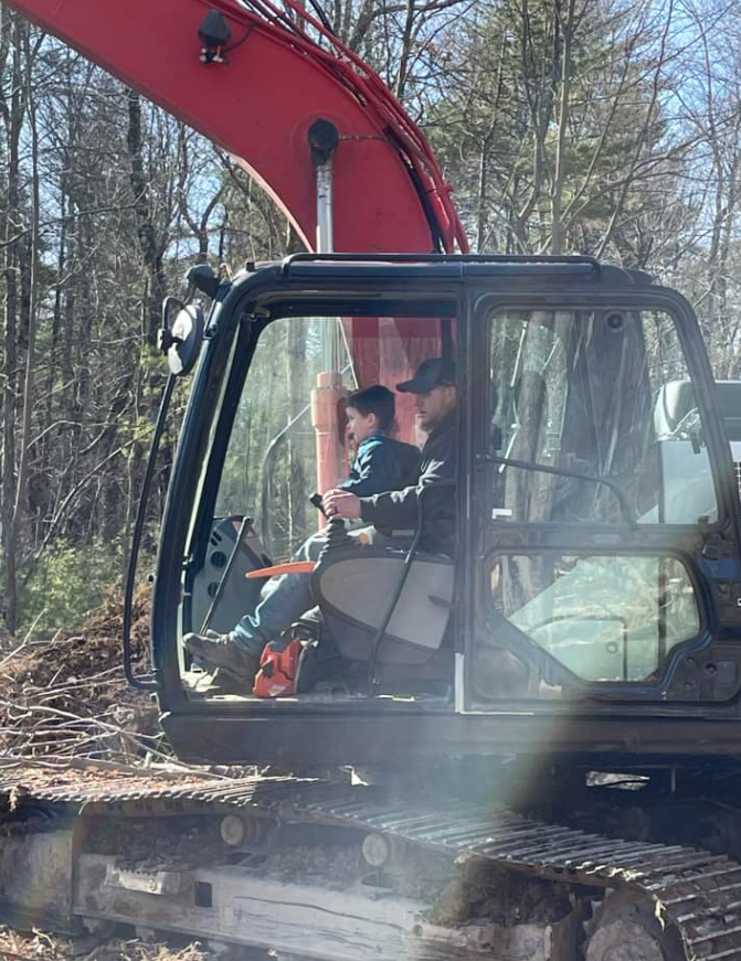 Man and child in excavator cab, operating controls, sunny outdoor setting.