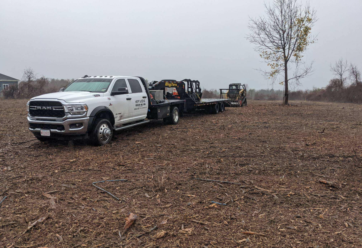 White truck pulling a flatbed trailer with a skid steer on a field of debris. Overcast day.