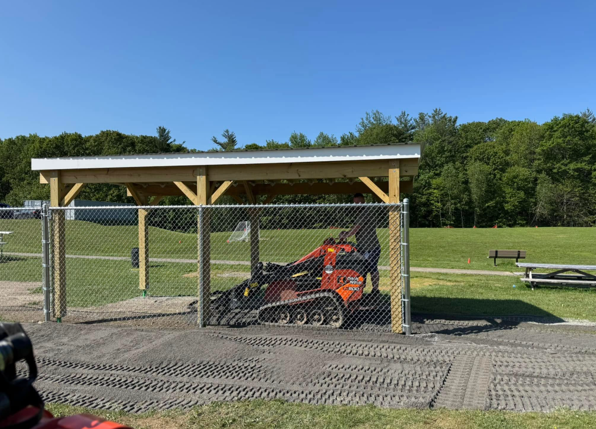 Orange trackhoe under a wooden shed and chain link fence on gravel; green trees and sky in background.