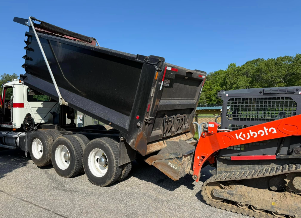 Dump truck with raised bed being loaded by an orange Kubota skid steer. Outdoors on gravel.
