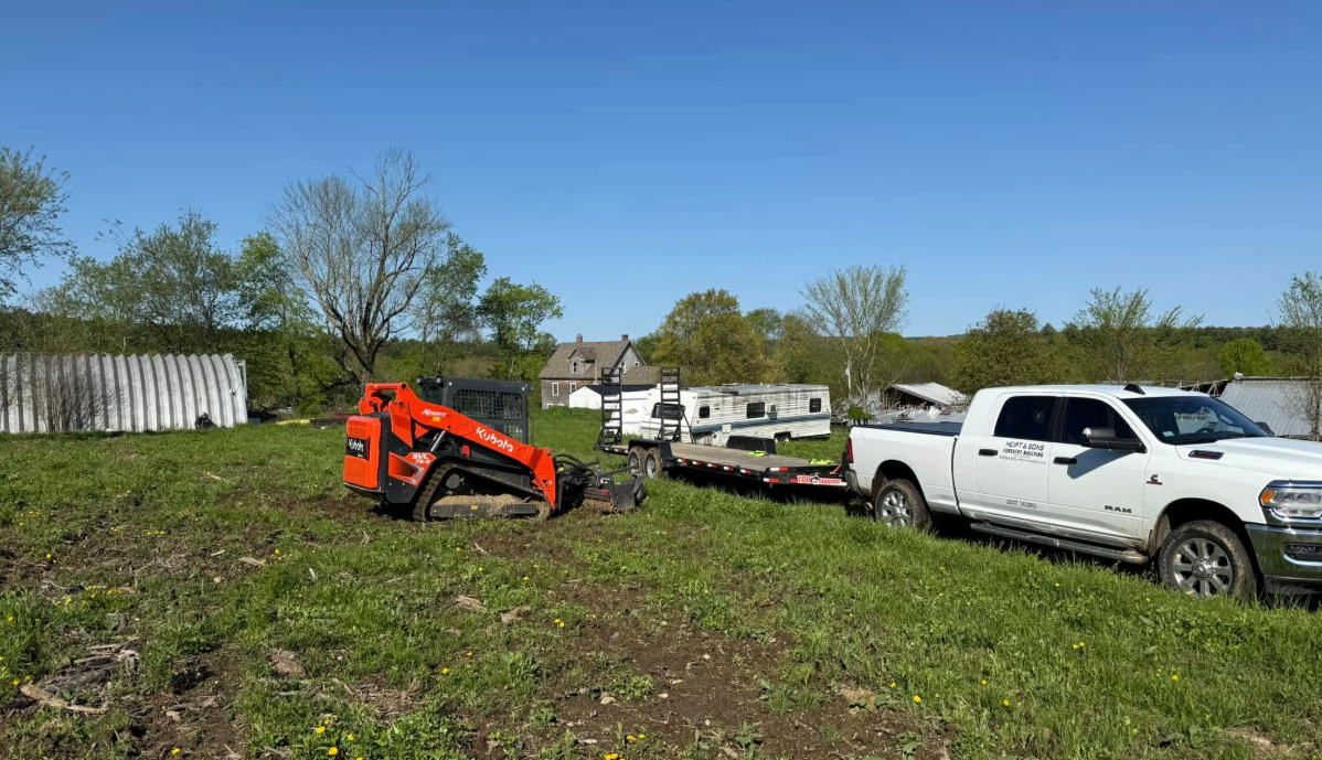 Orange skid steer, white pickup truck, and trailer on grassy field with a building.