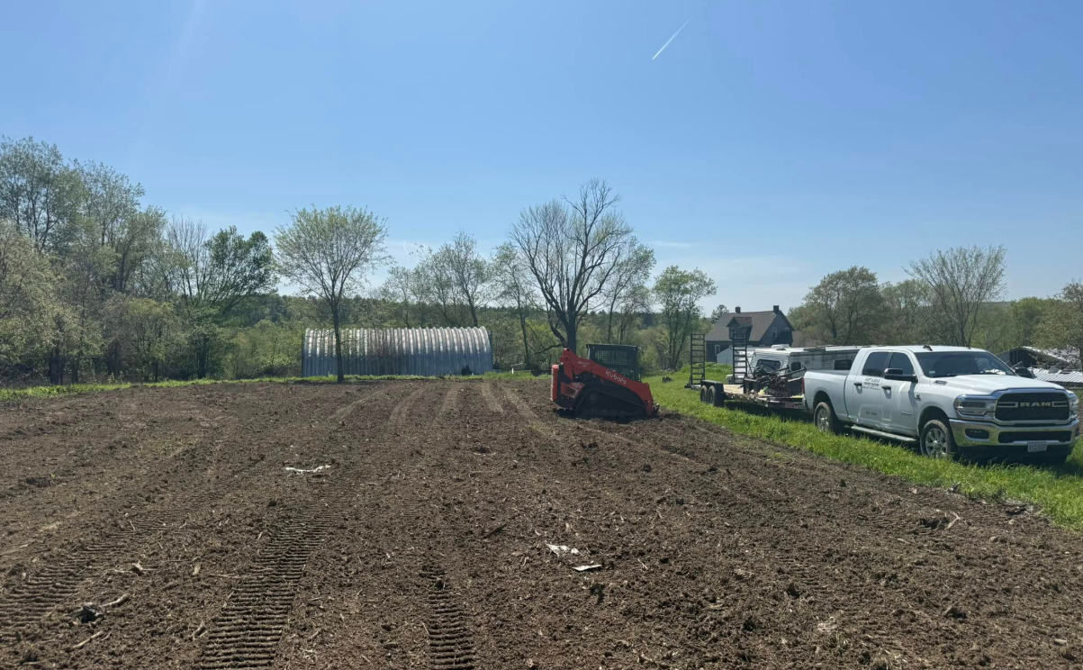 A plowed field with a tractor, truck, and trailer. Clear blue sky, green trees in background.