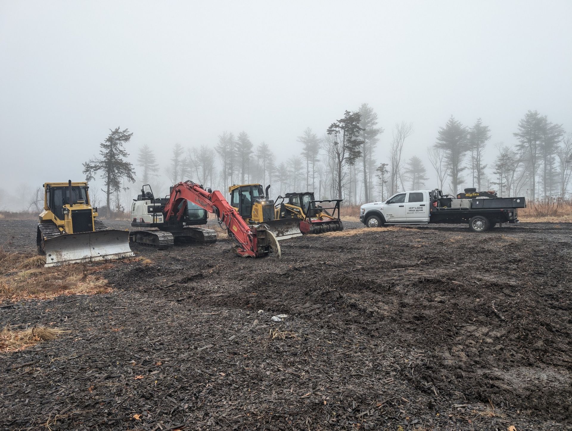 Construction equipment, including a bulldozer, excavators, and a flatbed truck, parked on a muddy site under foggy skies.