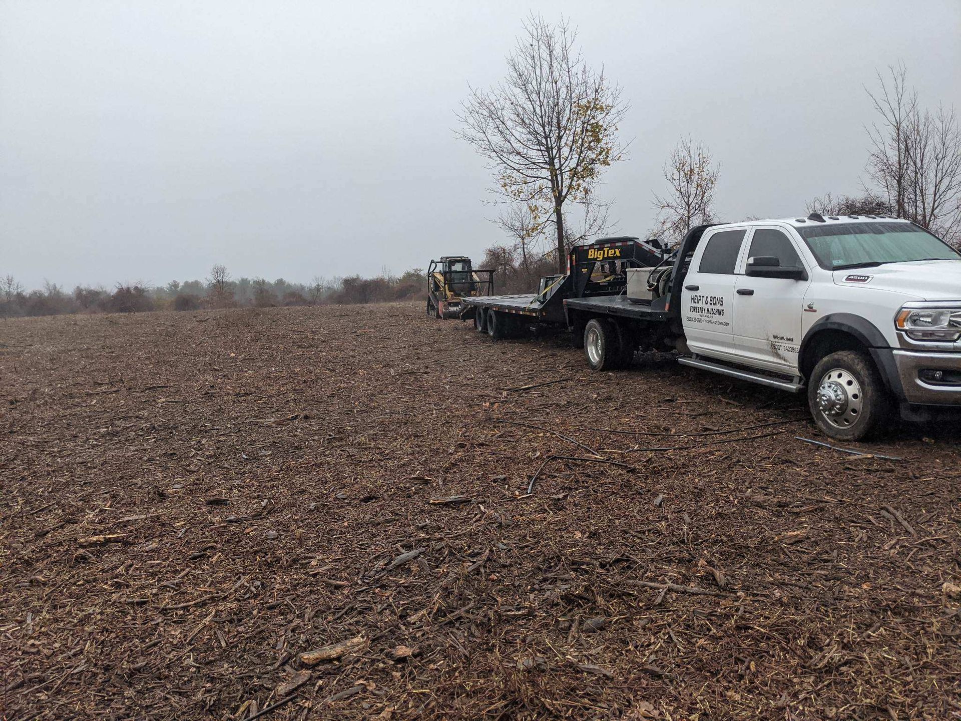 A white pickup truck with a trailer carrying construction equipment parked in a harvested field under a cloudy sky.