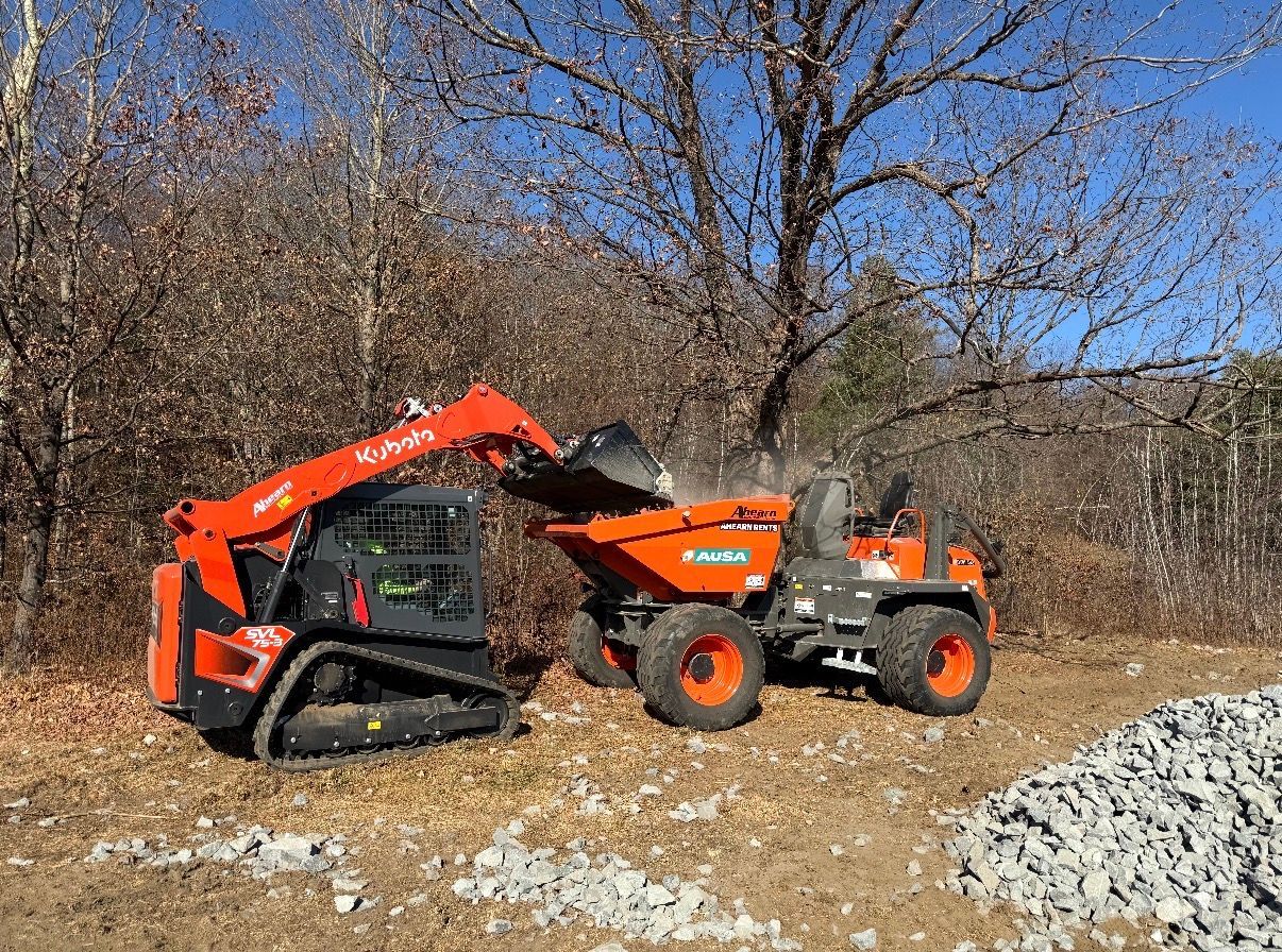 An orange Kubota skid-steer loader dumps dirt into an orange site dumper truck in a wooded area.