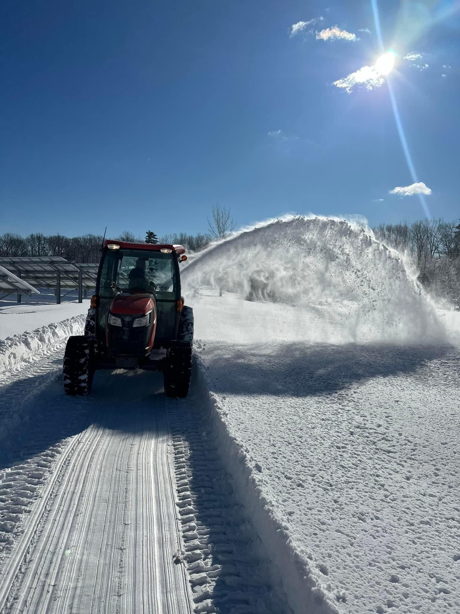A red tractor with a snowblower clears a path, casting a large plume of snow into the sunny, winter landscape.