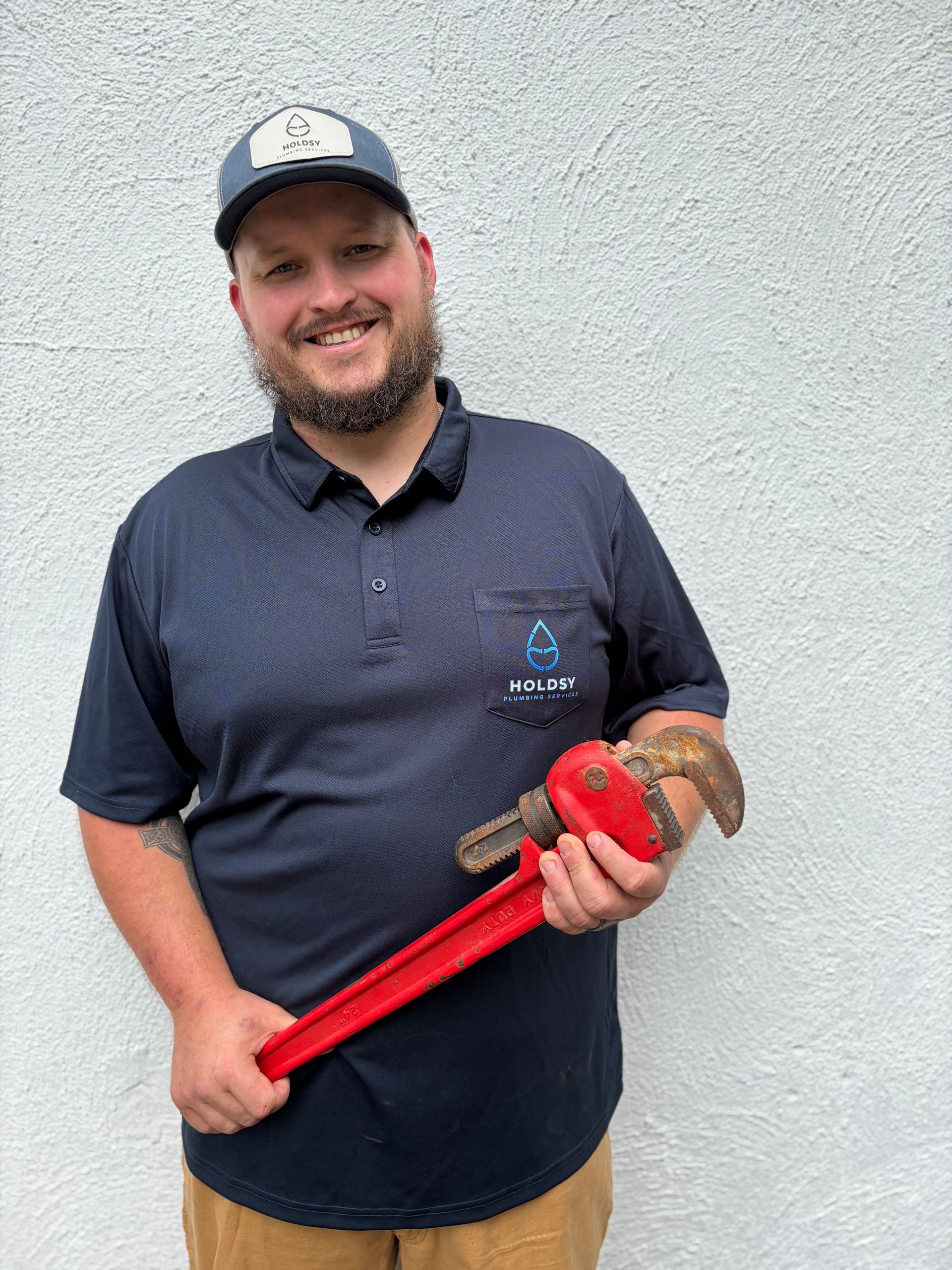 Smiling man in navy polo and cap holding a large red pipe wrench against a gray wall