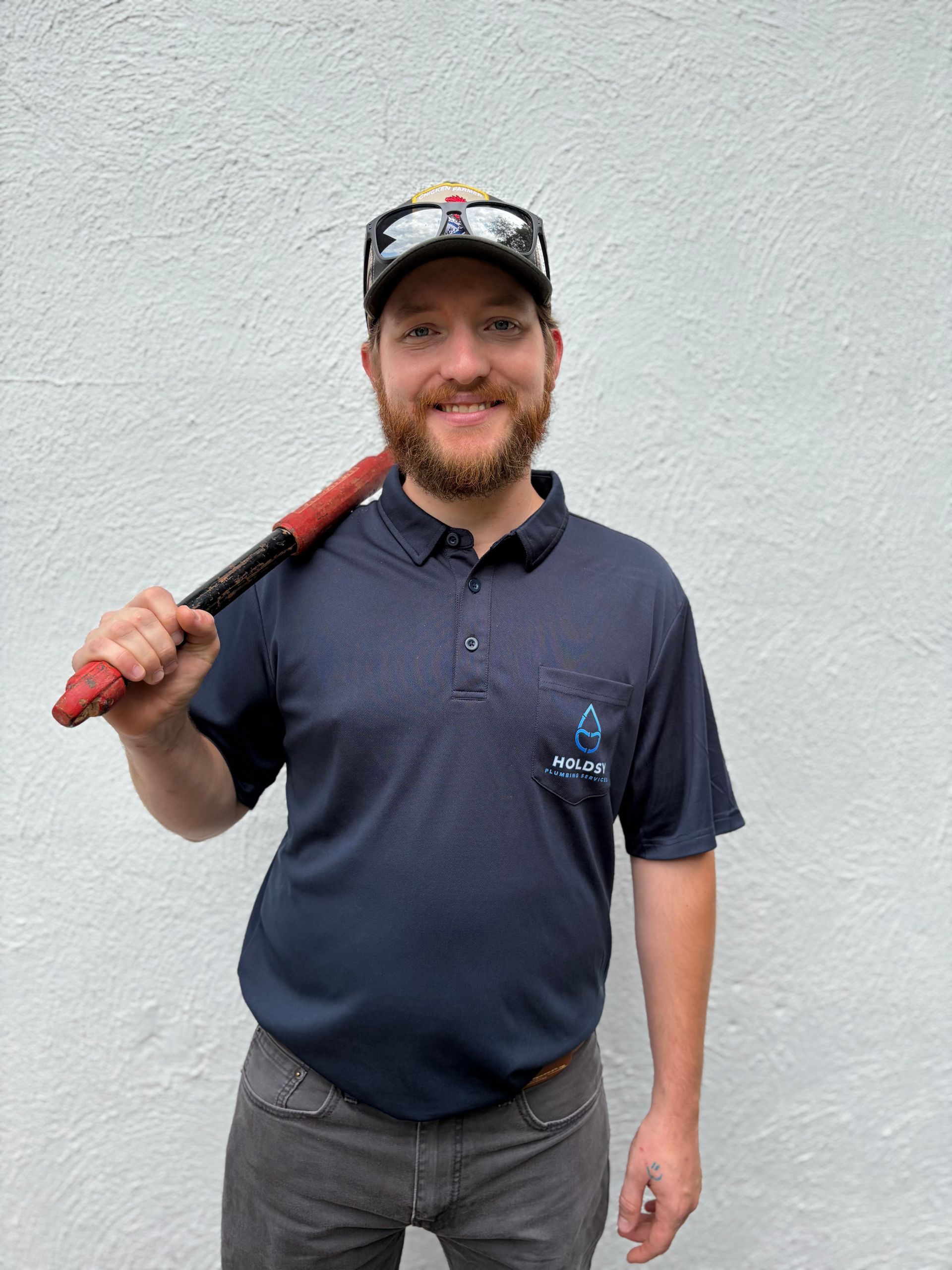 Person in navy polo and cap holding a red baseball bat over their shoulder against a white wall.