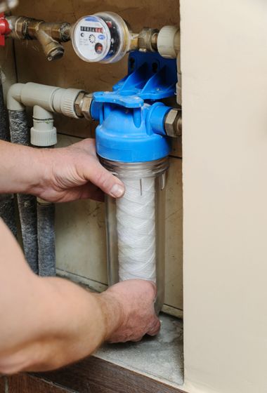 Hands installing a blue water filter cartridge under a sink, with pipes and a water meter visible.