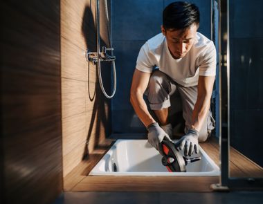 Person in white shirt using a jigsaw to cut a bathtub opening in a bathroom