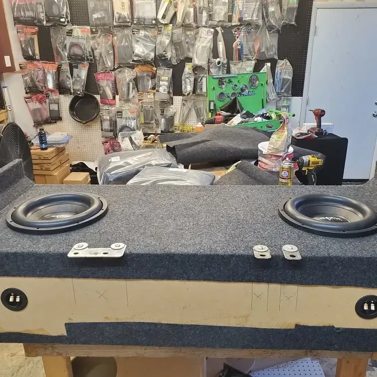 A subwoofer box with two speakers sits on a workbench in a cluttered shop. The box is covered in gray carpet.