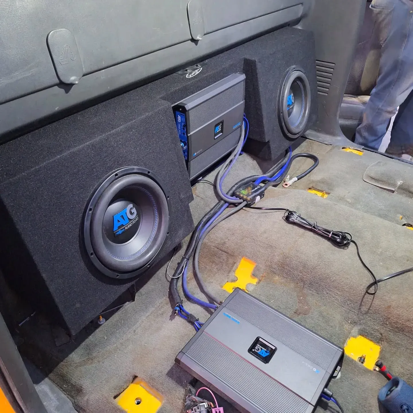 Two subwoofer boxes and a car amplifier setup in a vehicle interior, with exposed wiring on the floor.