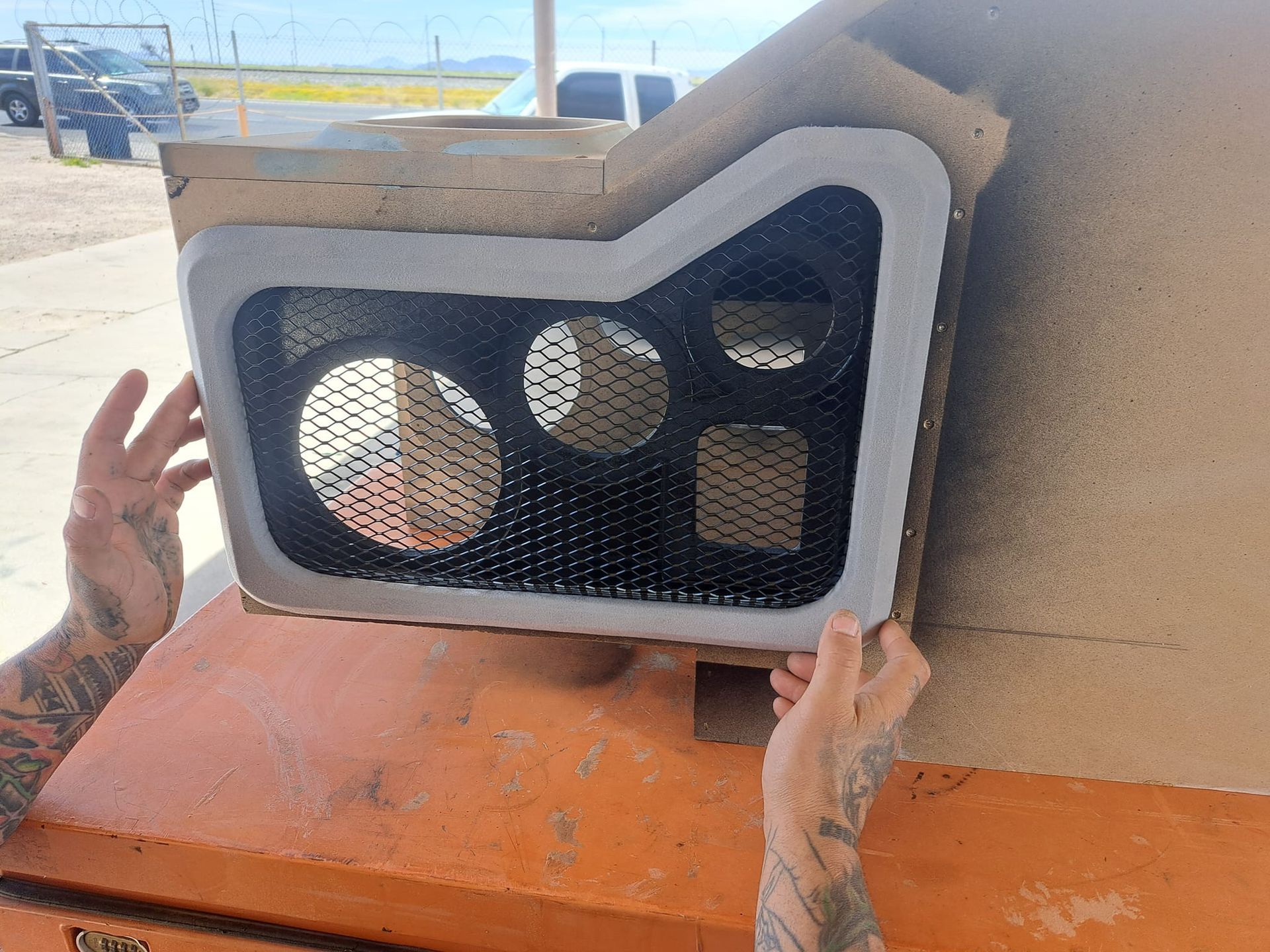 Person holds a custom speaker box with mesh over openings. Black, grey, and orange colors are prominent.