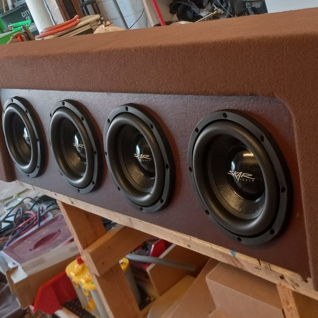 Four black subwoofers mounted in a brown speaker box, likely for a car. Box is on a wooden surface, garage setting.