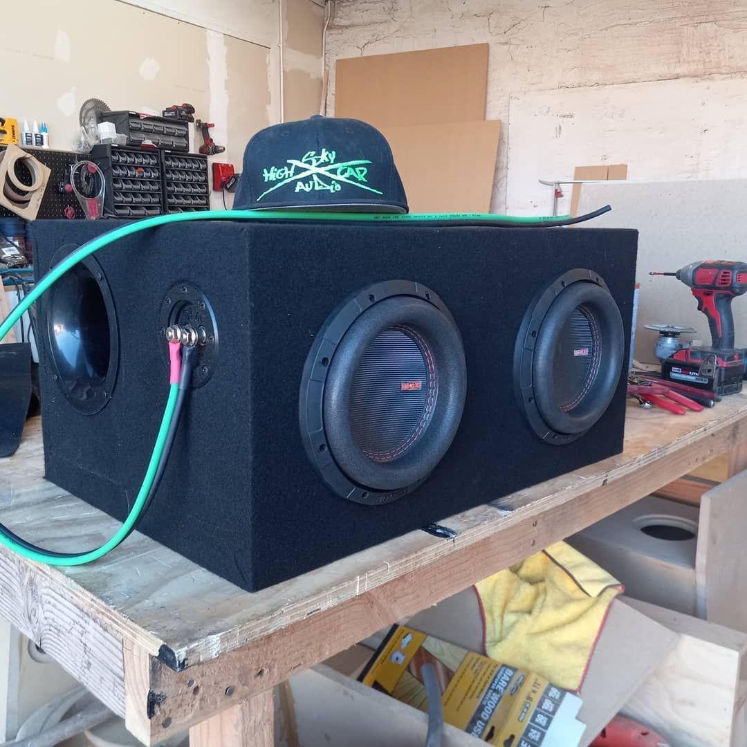 A black subwoofer box with two speakers and a port sits on a workbench. A baseball cap rests on top.