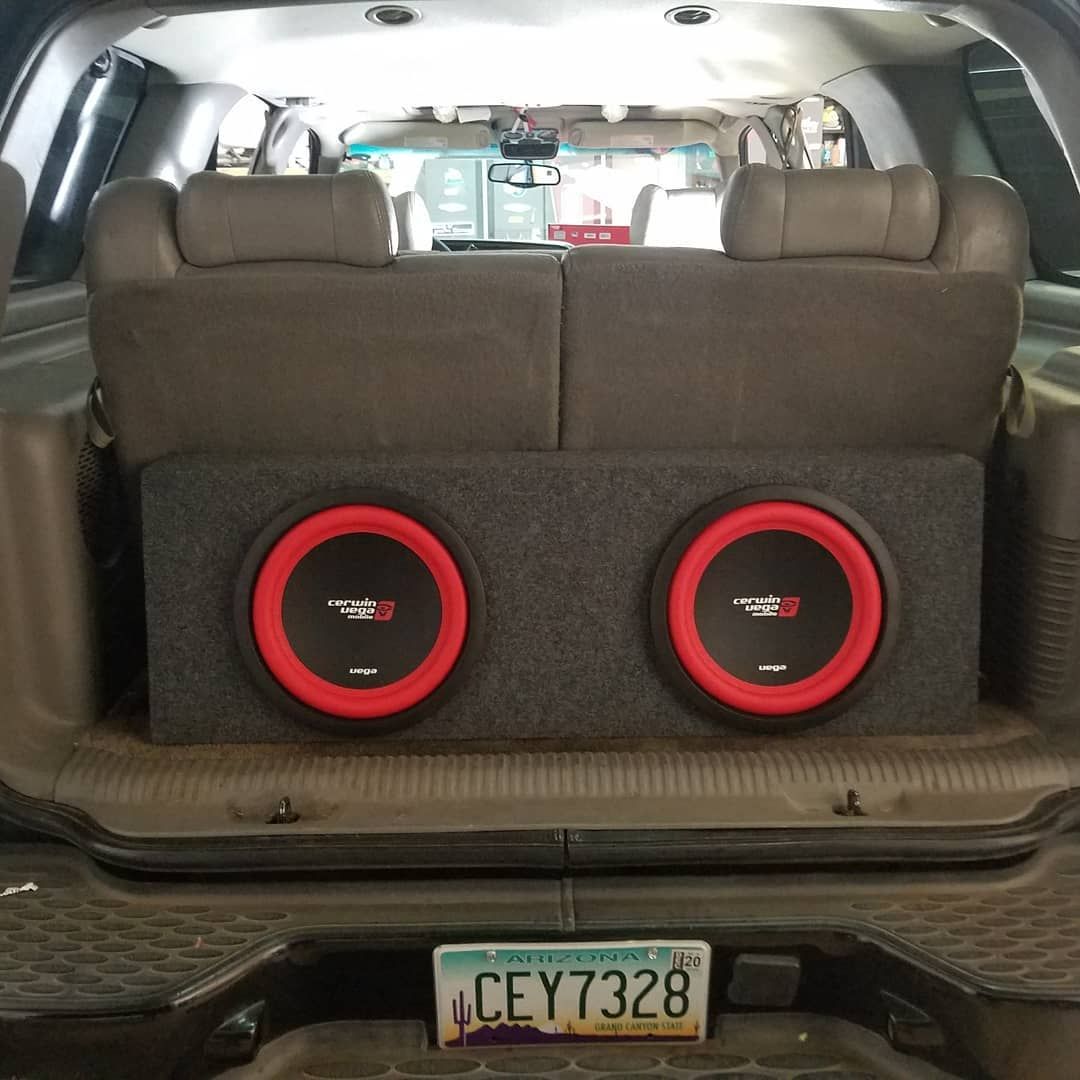 Two red-rimmed subwoofers in a gray box installed in the trunk of a vehicle, with a license plate visible.