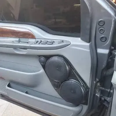 Car door with custom black speaker panel holding two speakers. The door is gray with wood trim and power window controls.