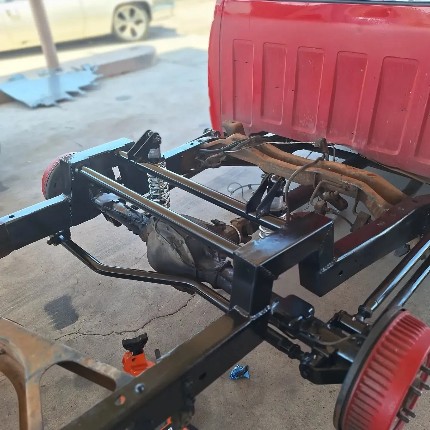 Red pickup truck frame in a garage, painted black. Rear suspension visible, with red brake drums.