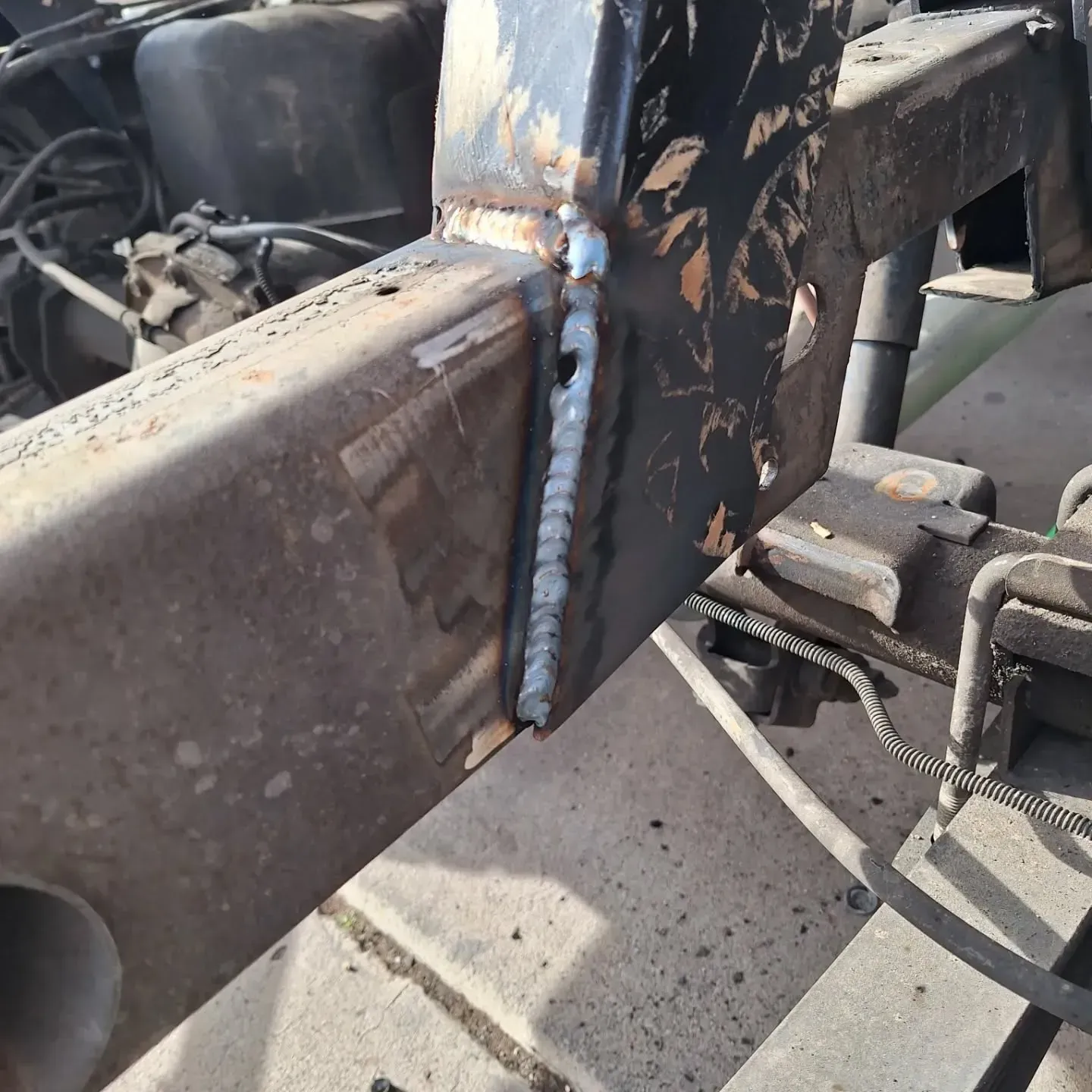 Close-up of a dark-colored metal frame with a weld joint. A vertical weld joins a rectangular beam to a flat plate.