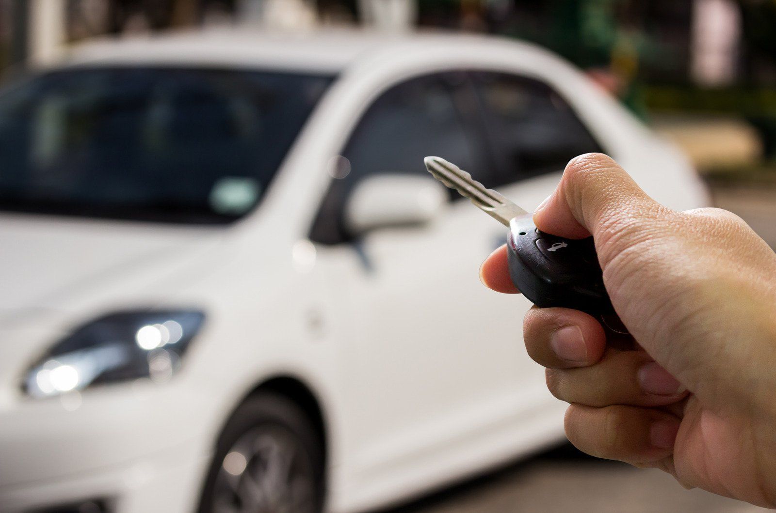 A hand holding a car key in front of a white sedan. The car is out of focus in the background.