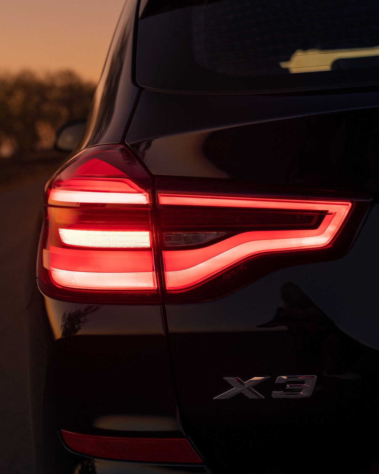 Rear view of a black BMW X3 SUV with illuminated red tail lights. Dusk setting with blurred background.