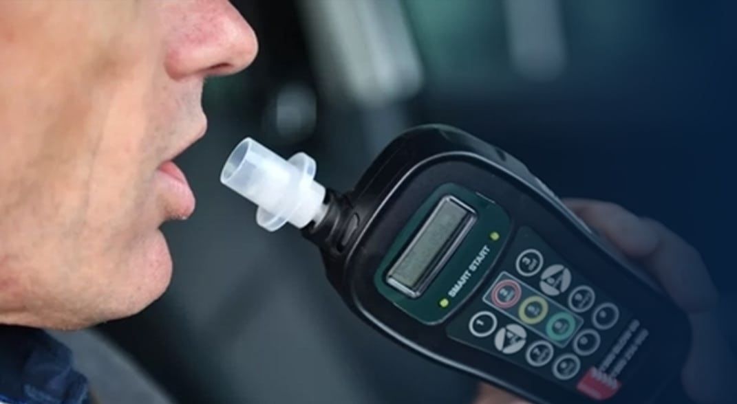 Man blowing into a handheld breathalyzer in a car. Device is black with a screen and buttons.