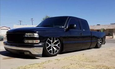 Black lowered pickup truck with chrome bumper and large silver wheels, parked on a sunny street.