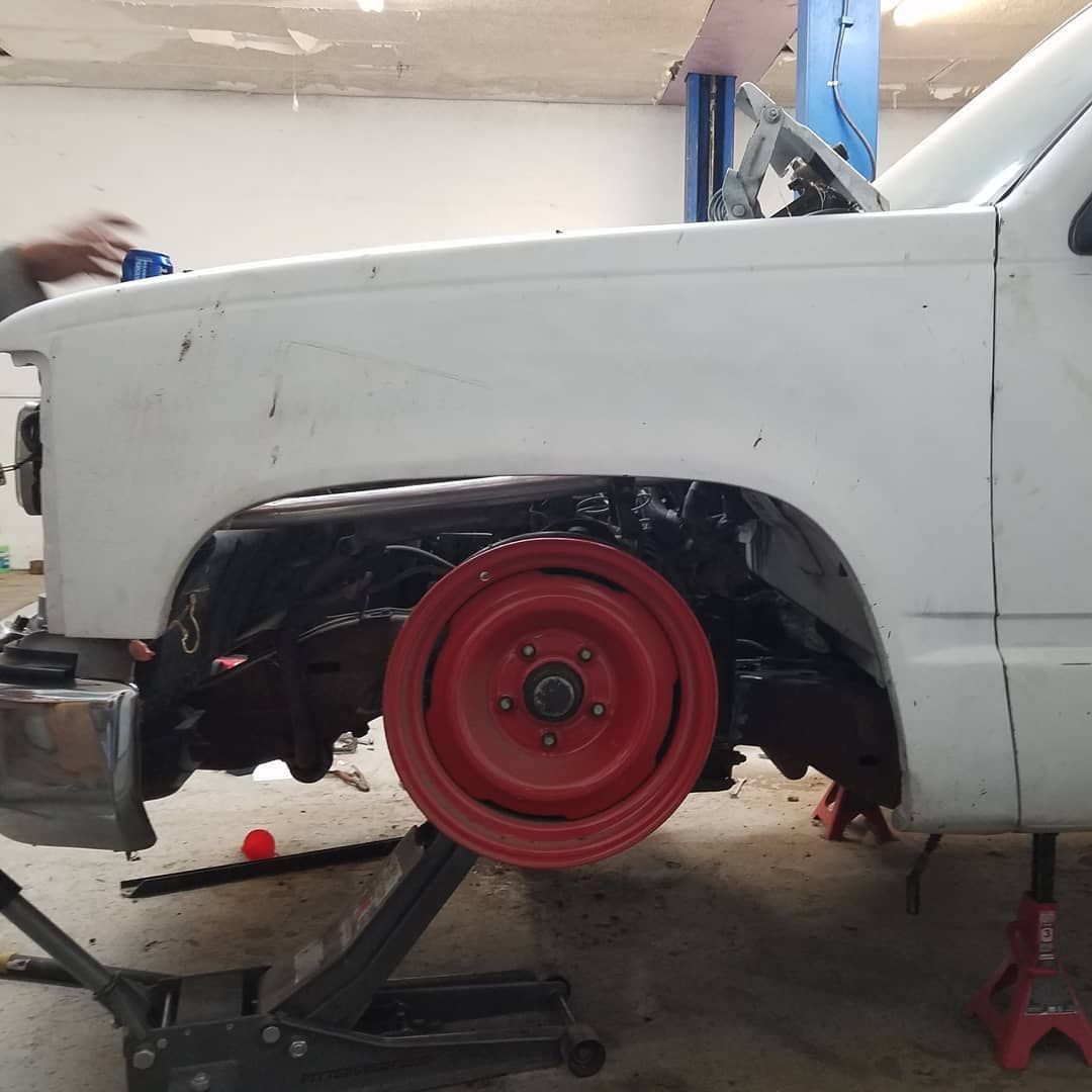 A white pickup truck with a red wheel, propped up on a jack in a garage. Someone is working on the truck.