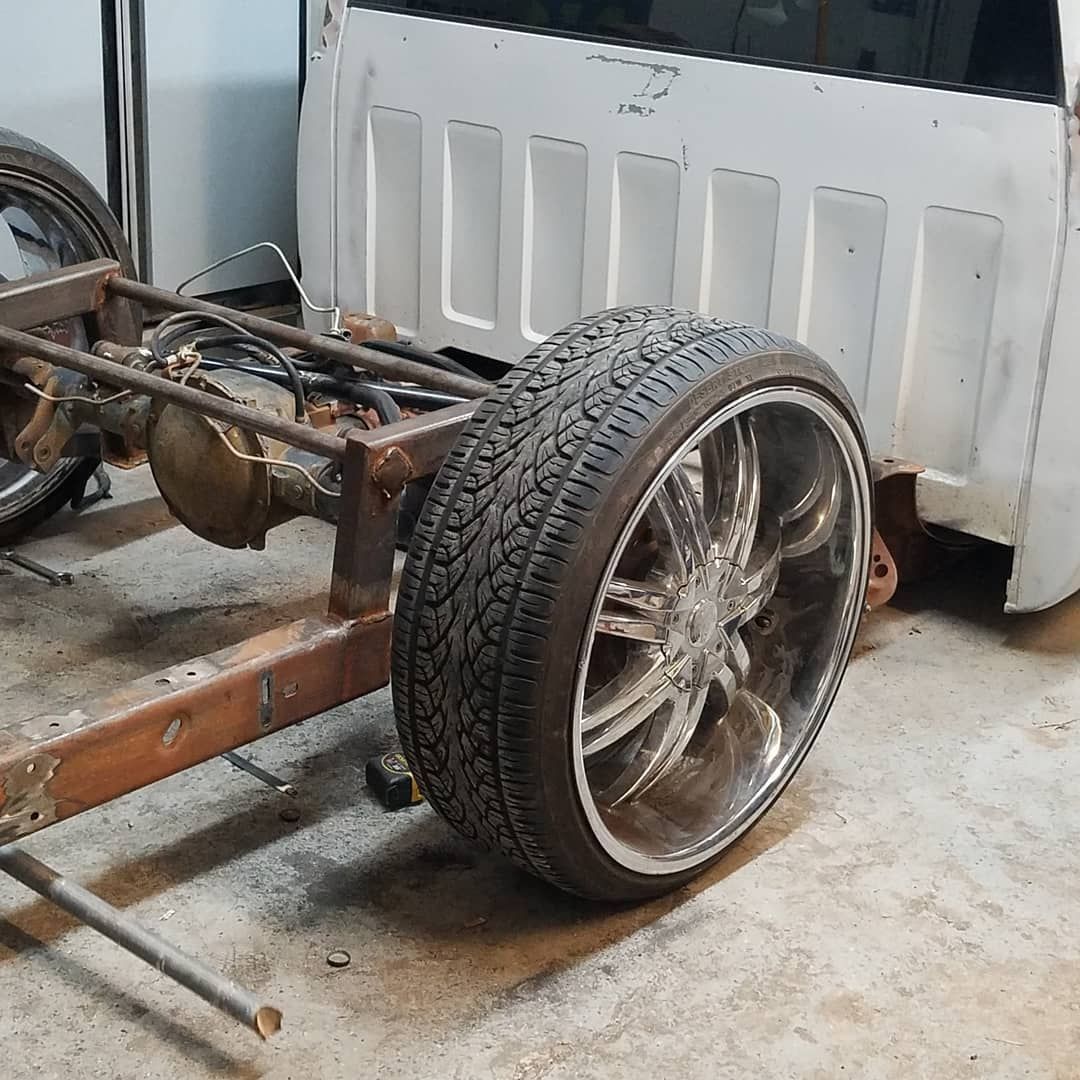 A pickup truck's chassis with a large chrome wheel and tire, the bed is white and unfinished.