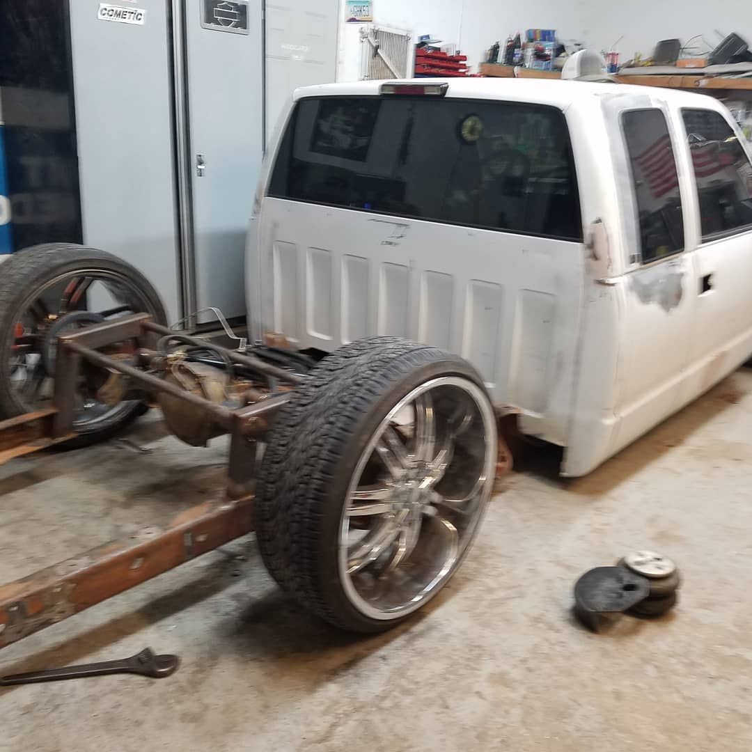 White pickup truck cab on a bare frame in a garage, showing large chrome wheels.