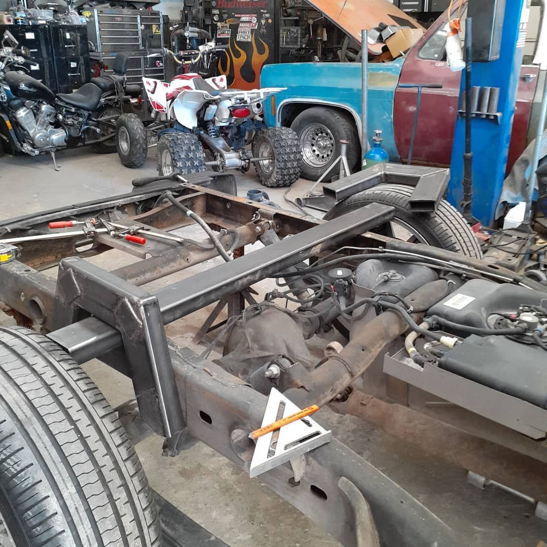 Truck frame with metal fabrication in a cluttered garage. Blue, red, and black vehicles are visible.