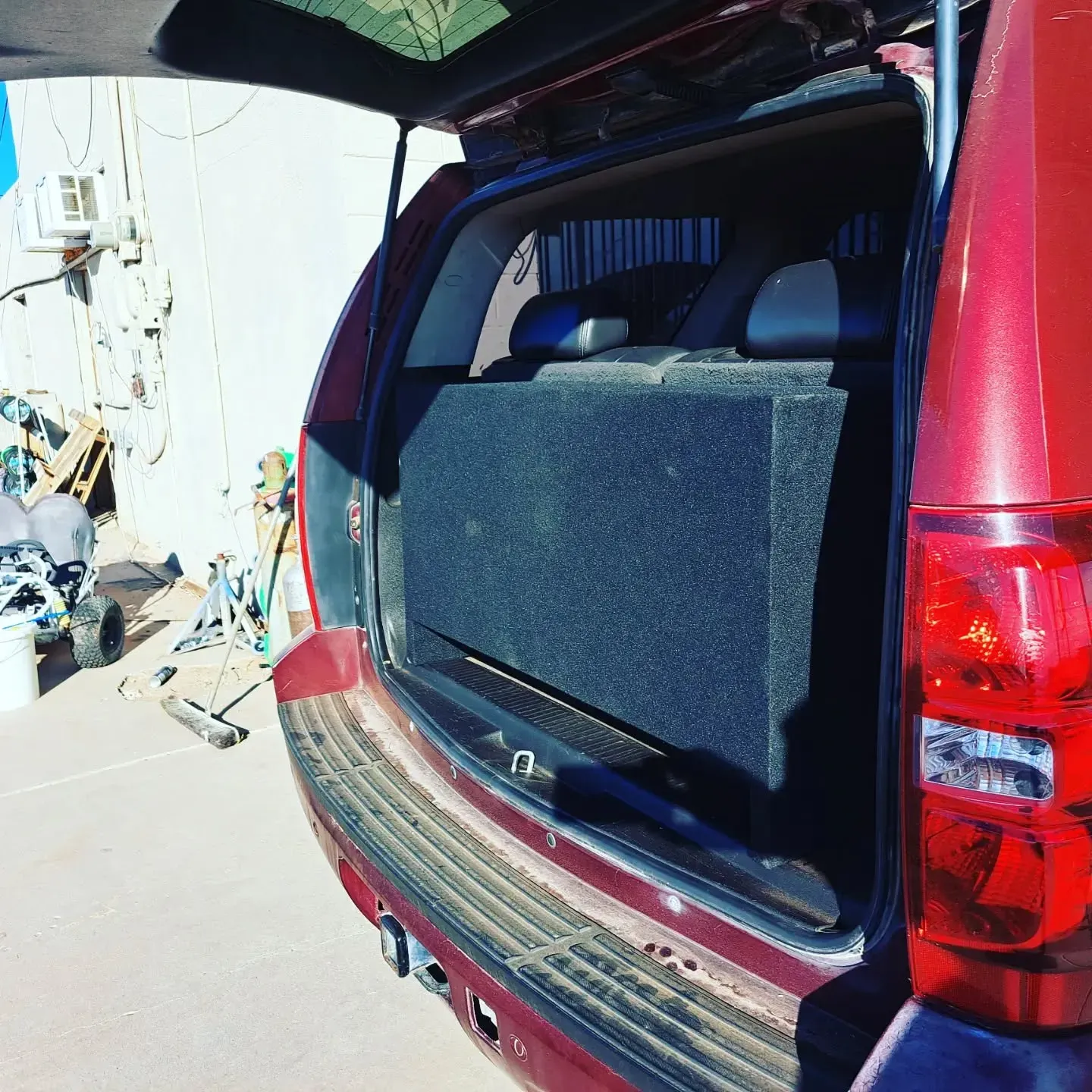 Open car trunk with a large black subwoofer box installed. Red car, outdoors.