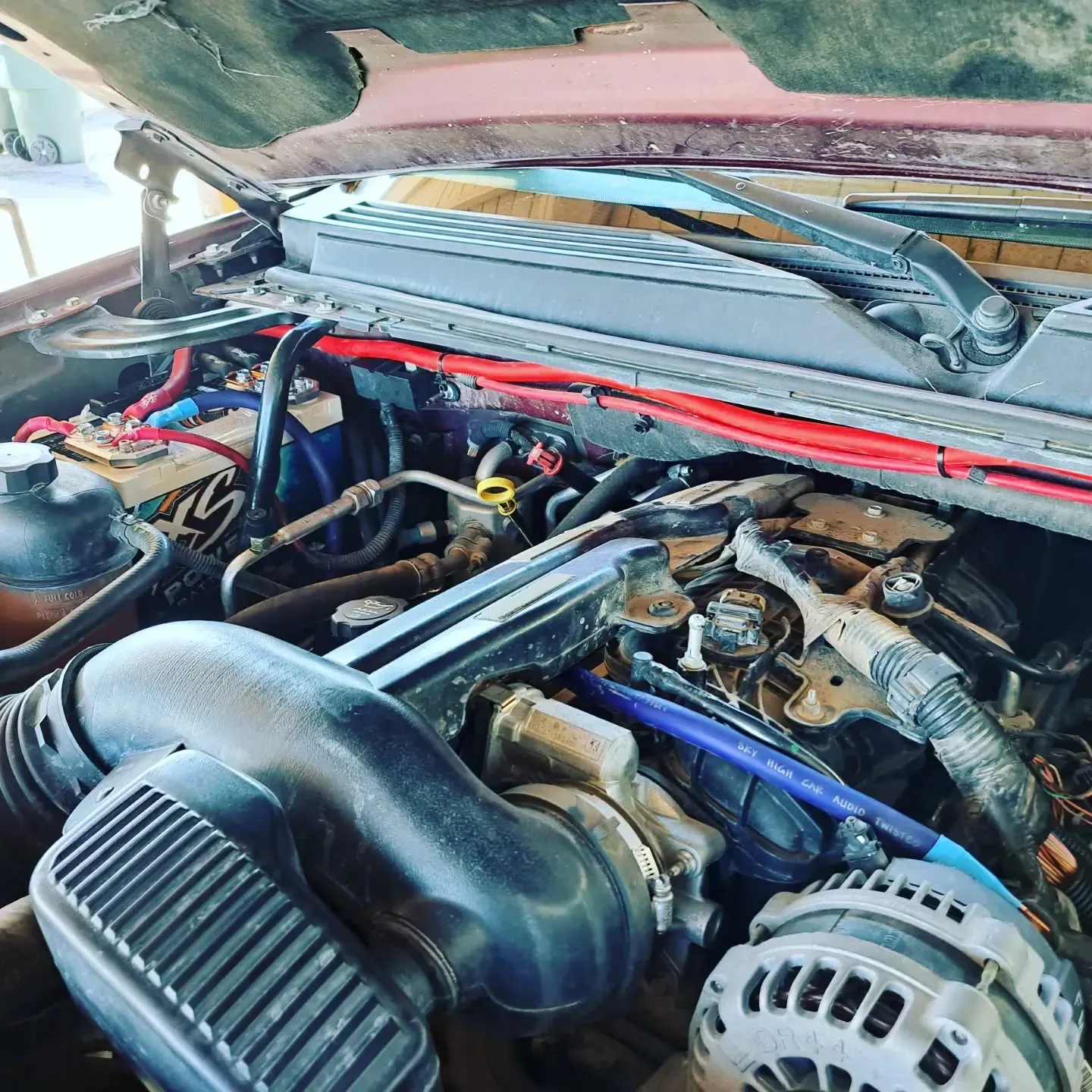 Open car engine bay, with hoses, wires, and various components visible. Red wires are prominent, with a black air intake.