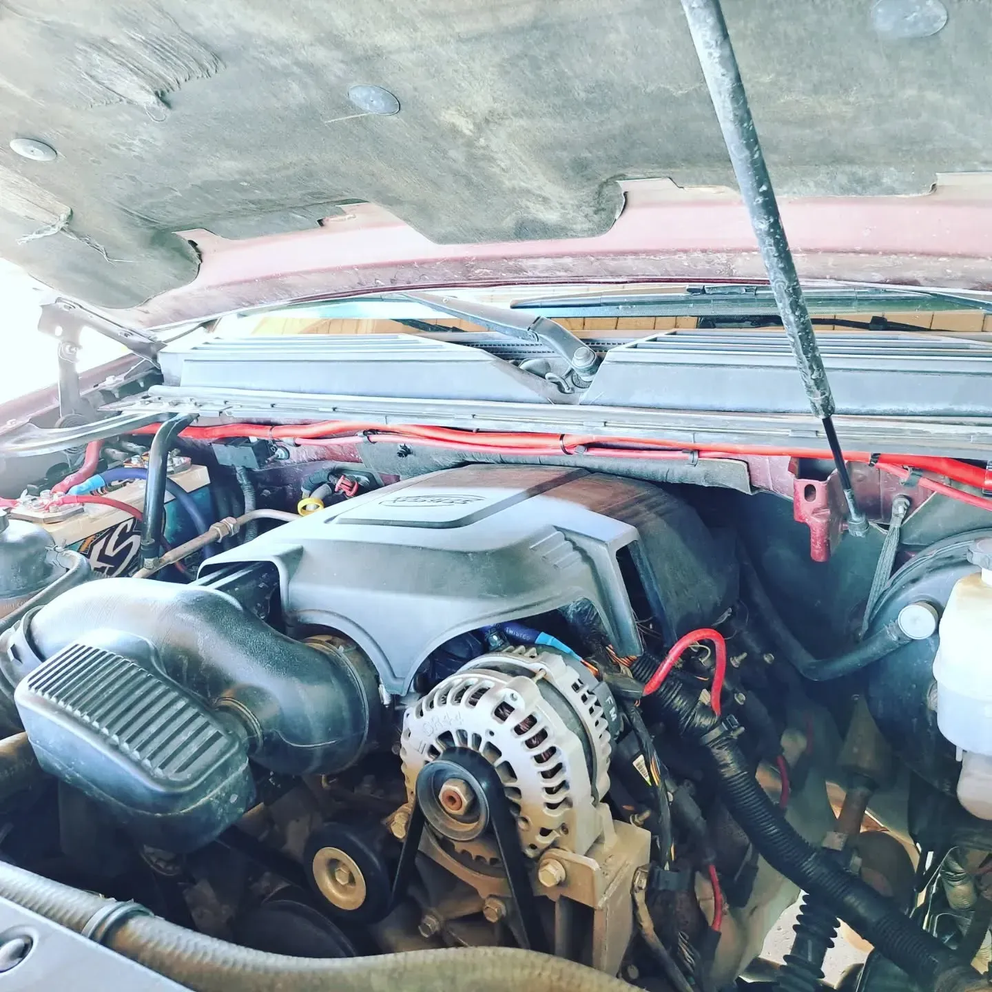 A vehicle engine bay with the hood open. The engine is black with silver components and red wiring, under a red hood.