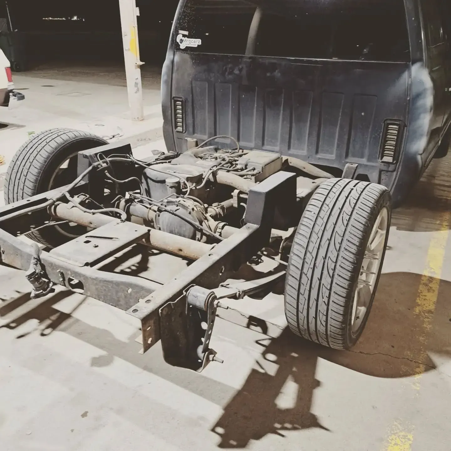 A black truck chassis and cab with exposed engine and rear tires in a dimly lit parking area.
