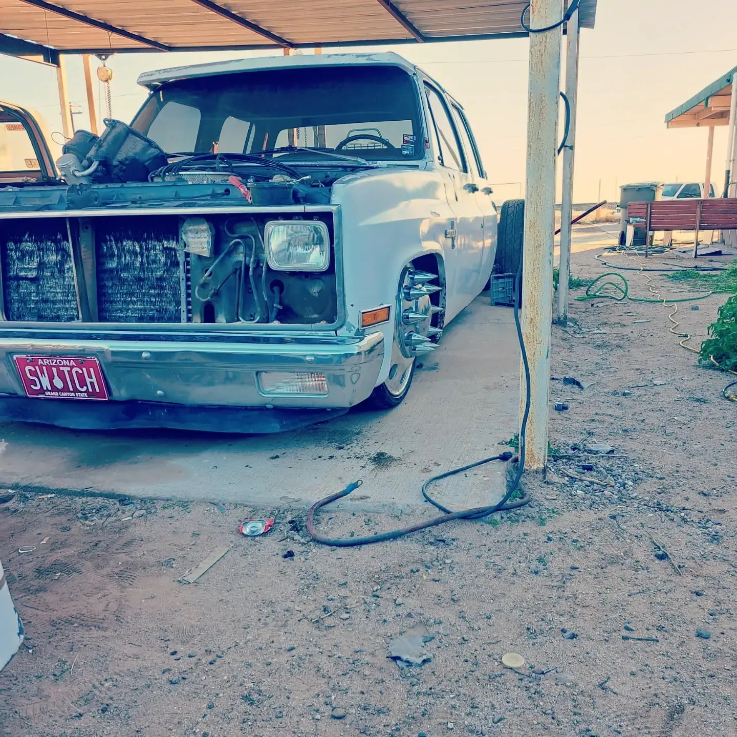 A lowrider truck with the hood open sits under a canopy in a desert sett